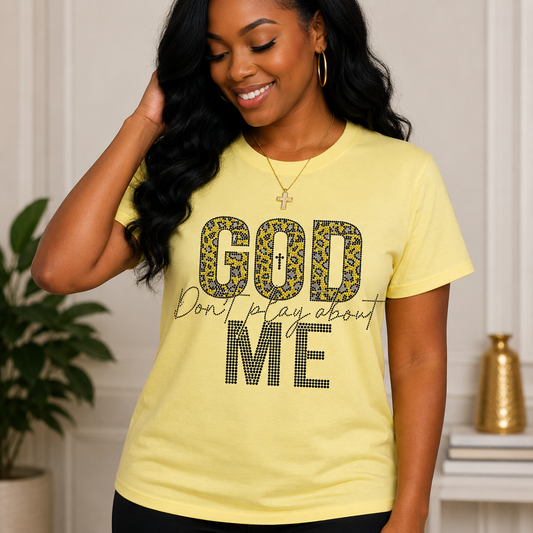 A woman wearing a pale yellow graphic T-shirt with the words 'GOD' and 'ME' in leopard-print and dotted lettering, with 'Don't play about' in script across the middle, posed in an indoor setting.