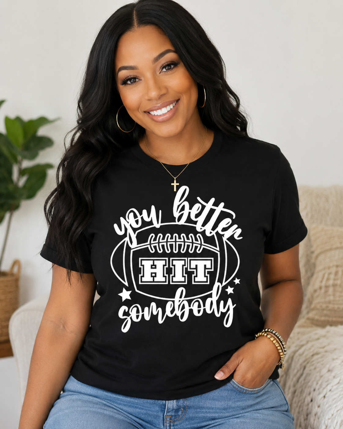 A woman wearing a black short-sleeve graphic T-shirt with a white football design and the text 'you better HIT somebody', paired with light blue jeans in a cozy indoor setting.
