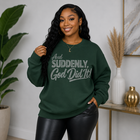 A woman modeling a dark green crewneck sweatshirt with the text 'And SUDDENLY, God Did It!' in a silver glitter-style font, paired with black faux-leather pants in an indoor styled setting.