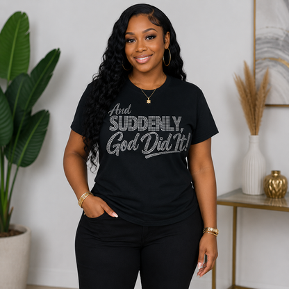 A woman wearing a black graphic t-shirt with the text 'And SUDDENLY, God Did It!' in a silver glitter-style font, paired with black pants, standing in a styled indoor room with a potted plant and a side table.