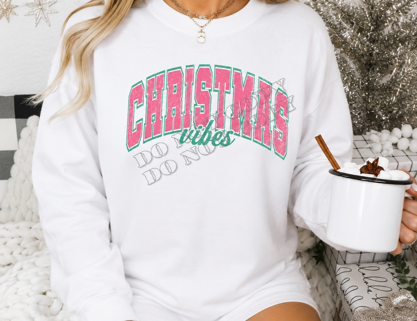 A woman wearing a white crewneck sweatshirt with a festive graphic that reads 'CHRISTMAS vibes DO NOT WORRY' in pink and green lettering. She is holding a white enamel mug with a cinnamon stick and hot chocolate, set against a cozy holiday background with a tinsel tree and knit blanket.