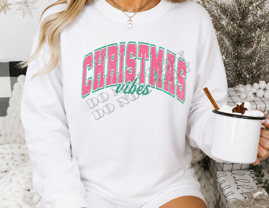 A woman wearing a white crewneck sweatshirt with a festive graphic that reads 'CHRISTMAS vibes DO NOT WORRY' in pink and green lettering. She is holding a white enamel mug with a cinnamon stick and hot chocolate, set against a cozy holiday background with a tinsel tree and knit blanket.