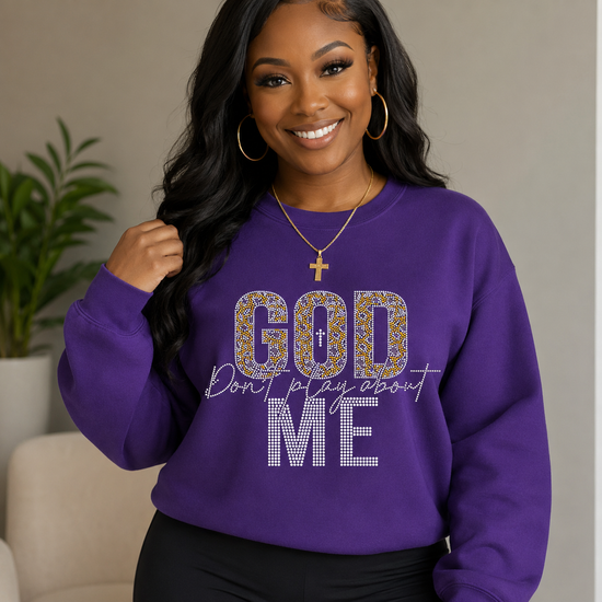 A woman wearing a purple crewneck sweatshirt with 'GOD Don't play about ME' printed on the front, styled with black pants, gold hoop earrings, and a gold cross necklace in an indoor setting.
