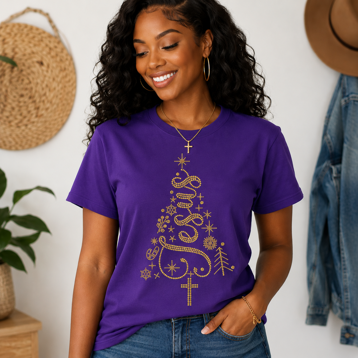 A woman wearing a purple short-sleeve t-shirt with a gold glittery Christmas tree graphic spelling 'Jesus' and stars, paired with blue jeans in a styled indoor setting.