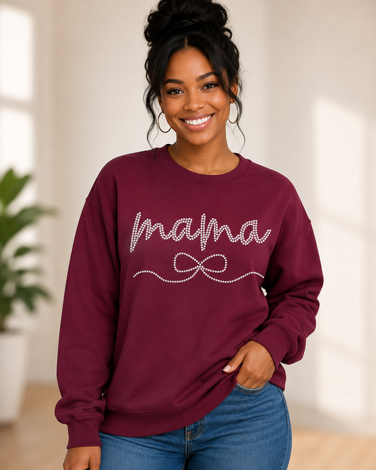 A woman wearing a burgundy crewneck sweatshirt with 'mama' and a bow design outlined in rhinestones, paired with blue jeans in a brightly lit indoor setting.