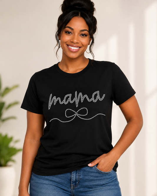 A woman wearing a black short-sleeve t-shirt with 'mama' and a bow design outlined in sparkling rhinestones, paired with blue jeans, set against a softly lit indoor background.