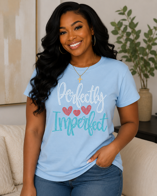 A smiling woman wearing a light blue graphic t-shirt with the text 'Perfectly Imperfect' in white and teal script, accented by three red hearts. She is posing in a cozy living room setting.
