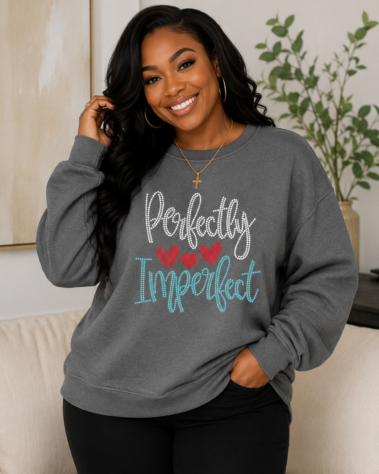 A woman wearing a heather gray sweatshirt with the text 'Perfectly Imperfect' in white and teal script, accented by three red hearts, posing in a cozy indoor setting.