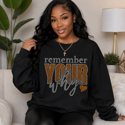 A woman wearing a black sweatshirt with a rhinestone-style graphic that reads 'remember YOUR why' in white, leopard-print brown, and silver, sitting on a light-colored couch in a living room setting.