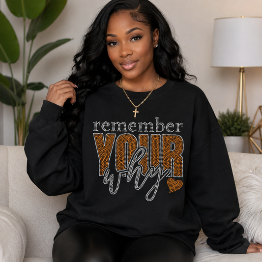A woman wearing a black sweatshirt with a rhinestone-style graphic that reads 'remember YOUR why' in white, leopard-print brown, and silver, sitting on a light-colored couch in a living room setting.