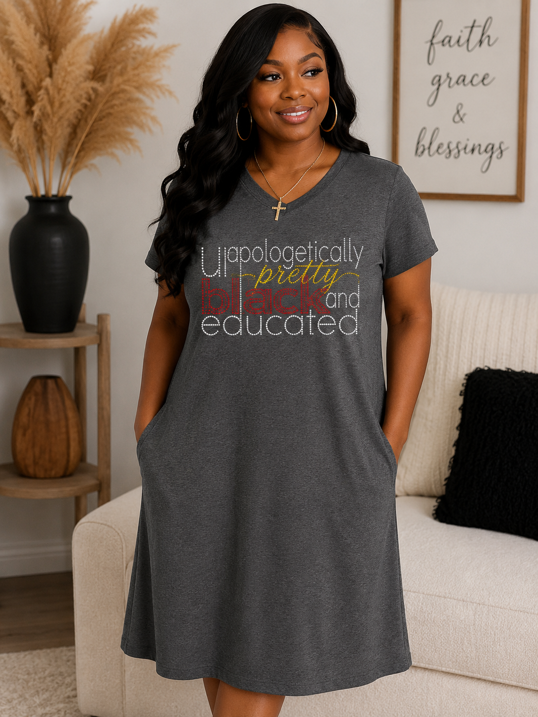 A woman wearing a dark gray heathered t-shirt dress with a colorful graphic text print that reads 'Unapologetically pretty, educated, and blessed'. She is standing in a living room setting with a beige couch, a black vase with dried pampas grass, and a framed print on the wall.