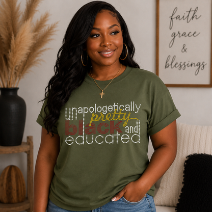 A woman wearing an olive green graphic t-shirt with the text 'unapologetically pretty black and educated' in white, yellow, and red fonts. She is standing in a cozy living room setting with a framed 'faith grace & blessings' sign on the wall behind her.